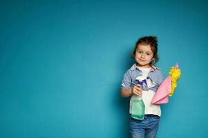 wenig Mädchen hält ein Staubtuch und ein sprühen mit Waschmittel im Hände. angenehm Reinigung Konzept. Blau Hintergrund. Kopieren Raum foto