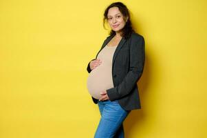 attraktiv Latina schwanger Frau im stilvoll Blazer, Bodysuit und Blau Jeans, lächelt beim Kamera, posiert mit Hände auf Bauch foto
