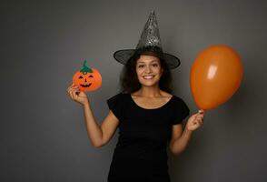heiter Frau im Magier Hut, gekleidet im schwarz , hält ein bunt Orange Luft Luftballons und zeigt an ein Filzschnitt handgemacht Kürbis, lächelt mit schön zahnig Lächeln suchen beim Kamera. Halloween Konzept foto