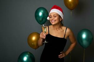 attraktiv Frau im Santa Hut und schwarz Kleid hat Spaß beim Weihnachten Party, lächelt zahnig Lächeln suchen beim Kamera halten Champagner Flöte, isoliert auf grau Hintergrund mit glänzend Gold Grün Luft Luftballons foto