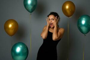 Porträt auf grau Mauer Hintergrund von ein erstaunt spanisch ziemlich Frau im schwarz Abend Kleid suchen beim Kamera mit Hände auf Gesicht, gegen grau Hintergrund mit Luxus Luft Luftballons. Feier Konzept foto