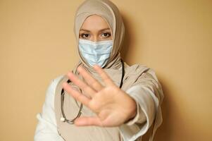 Porträt von Muslim Frau Arzt im Hijab und medizinisch Maske gestikulieren halt mit ihr Hand. Beige Hintergrund, Kopieren Raum. foto