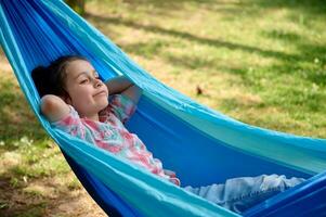 Nahansicht bezaubernd lächelnd Kind Mädchen entspannend auf Blau Hängematte im das Hinterhof oder Sommer- Lager, genießen ein Wochenende draußen foto