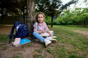 voll Länge Porträt Clever Schule Mädchen 6 Jahre alt, lächelt suchen beim Kamera, Sitzung auf Grün Gras und tun Hausaufgaben. foto