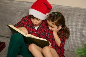 bezaubernd kaukasisch Kinder , Bruder im Santa's Hut und seine jünger Schwester Sitzung auf das Couch und lesen ein Buch, Fee Erzählungen auf das Weihnachten Nacht beim Zuhause foto