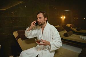 entspannt gut aussehend europäisch Mann im Weiß Bademantel halten ein Tasse von Tee und reden auf Zelle Telefon, Sitzung auf ein Chaise Salon während entspannend durch Whirlpool im Wellness Spa foto