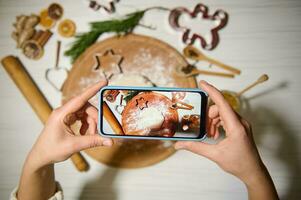 oben Aussicht von Hände halten Handy, Mobiltelefon Telefon und Herstellung Foto von ein Lebkuchen Teig auf ein runden hölzern Tafel Nächster zu ein rollen Stift und Küche Utensilien zum Herstellung Weihnachten Kekse. Telefon im Leben Aussicht Modus