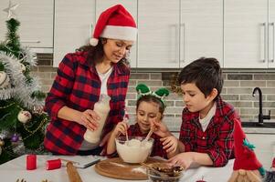 glücklich jung liebend Mutter im kariert Hemd und Santa Hut gießt etwas Milch in Schüssel mit Mehl und ihr bezaubernd Kinder- süß Mädchen und gut aussehend Junge Mischen und kneten trocken Zutaten, Herstellung Teig foto