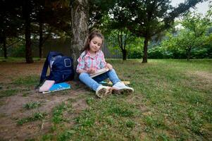 voll Länge Porträt bezaubernd Schule Mädchen 6 Jahre alt, Sitzung auf Grün Gras im ein Park, tun Hausaufgaben. zurück zu Schule foto