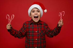 heiter komisch Kind Junge, schön Kind posiert gegen farbig rot Hintergrund mit Weihnachten Süßigkeiten Stöcke, zuckerhaltig gestreift Lutscher im Hände, freut sich suchen beim Kamera. Neu Jahr Konzept mit Kopieren Raum foto