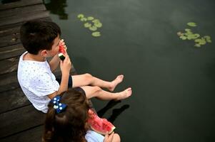 Kinder mit Wassermelone ruhen auf ein Seebrücke und bewundern Wasser Lilien wachsend unter Fluss Wasser. Landschaft Sommer- Urlaube Konzept. Junge und Mädchen haben Spaß draußen foto