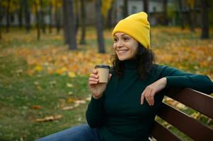 Erfreut jung Frau im warm Wolle Gelb gestrickt Hut lächelt zahnig Lächeln Sitzung auf ein hölzern Bank mit ein wegbringen Kunst Papier Tasse, ruhen im ein herbstlich Wald Park mit Ahorn Bäume auf Hintergrund foto