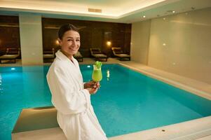 charmant Frau im Weiß Bademantel steht durch das Schwimmbad im luxuriös ein Wellness Spa Komplex, hält frisch gedrückt Vitamin Saft im Hände, lächelt suchen beim Kamera. gesund Lebensstil, Erholung Konzept foto