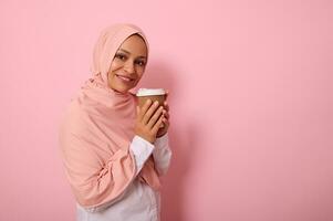 Arabisch Muslim ziemlich Frau mit bedeckt Kopf im Rosa Hijab Trinken heiß trinken, Tee oder Kaffee von Einweg Karton wegbringen Tasse, Stehen drei Viertel gegen farbig Hintergrund mit Kopieren Raum foto