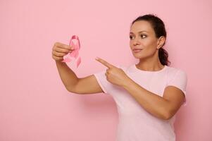 schön gemischt Rennen Frau tragen Rosa T-Shirt zeigen zu ein Rosa Satin- Band im ihr Hand, farbig Hintergrund Kopieren Raum. Konzept von 1 st Oktober, International Tag von Kampf Brust Krebs Krankheit foto