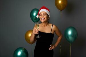 ziemlich Frau im Santa Hut und Abend Kleid hat Spaß beim Weihnachten Party, lächelt zahnig Lächeln suchen beim Kamera halten Champagner Flöte, isoliert auf grau Hintergrund mit glänzend Gold Grün Luft Luftballons foto