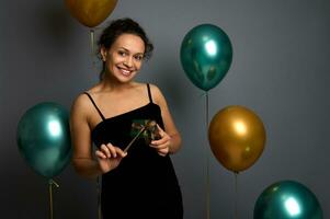 charmant schön jung Frau im schwarz Abend Kleid hält ein Weihnachten Geschenk hinter ein Gold Bogen und lächelt süß beim das Kamera, isoliert auf ein grau Hintergrund dekoriert mit Gold und Grün Luftballons foto