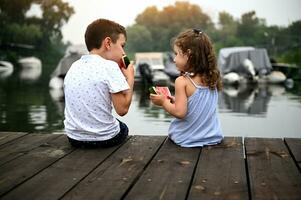 Junge und Mädchen Sitzung auf das Seebrücke beim Sonnenuntergang gegen das Hintergrund von Boote , Essen Wassermelone . Freundschaft, Zärtlichkeit, Familie Beziehungen Konzept. glücklich Kindheit. foto