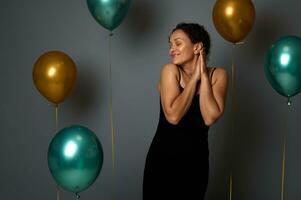 heiter ziemlich Frau posiert mit geschlossen Augen gegen grau Mauer Hintergrund mit schön aufgeblasen festlich golden und Grün metallisch Luft Luftballons auf Kopieren Raum zum Weihnachten Neu Jahr Jahrestag Anzeige foto