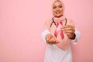 verschwommen Muslim Arzt im Rosa Hijab posieren auf Rosa Hintergrund, zeigen Unterstützung, Solidarität zu Patienten Krebs Überlebende. Welt Tag von Kampf Krebs. Fokus auf Rosa Brust Krebs Bewusstsein Band foto