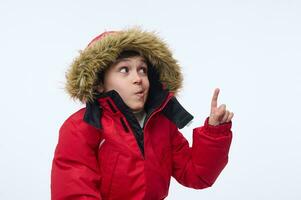 gut aussehend heiter vorpubertär, Schule alt europäisch Junge tragen hell rot Parka mit Haube, lächelnd zahnig Lächeln suchen beim Kamera und zeigen mit Finger auf ein Kopieren Raum von Weiß Hintergrund foto