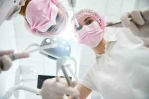 Unterseite Aussicht von Gruppe von Zahnarzt und Dental Assistent im Rosa schützend Maske halten Dental Werkzeuge und Instrumente zum Untersuchung und gelehnt gegenüber ein geduldig auf das Hintergrund von medizinisch Licht Lampe foto