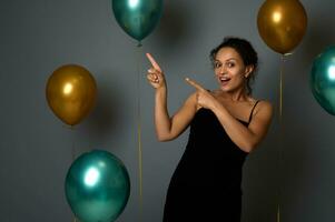 attraktiv froh Frau im Abend Kleidung sieht aus beim Kamera und Punkte auf ein grau Hintergrund Kopieren Raum dekoriert mit golden Grün Luft Luftballons zum Weihnachten, Jahrestag , Neu Jahr Werbung foto