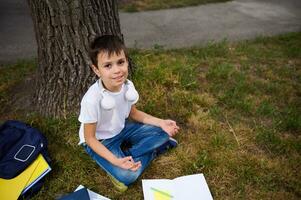gut aussehend Schule Kind Junge Sitzung im Lotus Position auf das Grün Gras von Öffentlichkeit Park und meditieren , suchen oben beim das Kamera, süß lächelnd. Arbeitsmappen und Schule liefert Lügen auf das Gras foto
