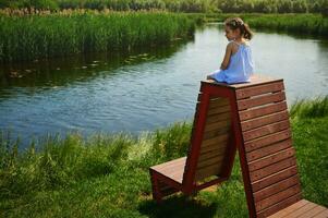 ein süß Mädchen im ein Blau Kleid und zwei Pferdeschwänze sitzt auf oben von ein hölzern Bank und glücklich Uhren ein schön Aussicht von das Fluss im das Landschaft foto