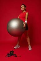voll Länge Porträt von ein schön aktiv Frau im Sportbekleidung ausüben mit passen Ball gegen rot farbig Hintergrund mit Kopieren Raum foto