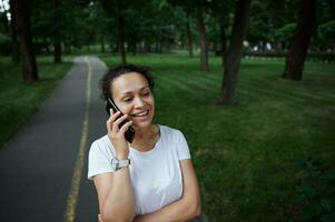 schön weiblich Tourist im Weiß T-Shirt, Gespräche auf Handy, Mobiltelefon Telefon während spazieren gehen das Gasse von ein Stadt Park Ö Sommer- Tag foto