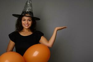 lächelnd süß Frau tragen Magie Halloween Hut sieht aus beim das Kamera und hält Orange Luftballons im einer Hand und Aufzüge das andere Palme hoch, halten imaginär Kopieren Raum zum Anzeige auf grau Hintergrund. Halloween foto