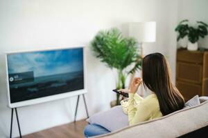 asiatisch Frau Aufpassen Clever Fernseher und mit Fernbedienung Regler Hand halten Fernsehen Audio- Fernbedienung Steuerung beim Zuhause mit das Fernbedienung Steuerung Fernsehen foto