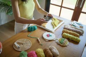 asiatisch Frau nehmen Smartphone Foto schlagen Nadel. Telefon Buchung auf Sozial Netzwerke im Studio Werkstatt. Designer Arbeitsplatz handgemacht Kunst Projekt DIY Stickerei