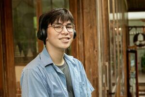 Porträt von asiatisch jung Erwachsene im Blau Shirt, tragen Brille, halten Kopfhörer und Stehen im Vorderseite von das Tür von Kaffee Geschäft, Konzept zum Lebensstil von Jugendliche um das Welt. foto