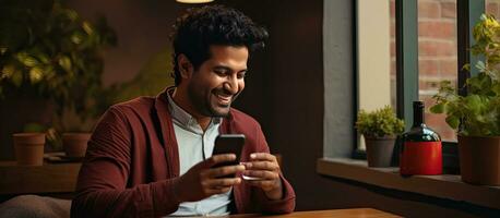 glücklich indisch tausendjährig Surfen Sozial Netzwerke auf Handy, Mobiltelefon Telefon und Trinken Kaffee beim Zuhause Kopieren Raum foto