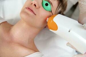 teilweise Aussicht von jung Frau mit schützend uv Brille Empfang Laser- Behandlung mit modern Laser- Gerät foto