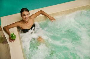 oben Aussicht von ein attraktiv Mitte alt europäisch Frau im schwarz Badeanzug halten ein frisch gedrückt Vitamin Saft und suchen beim Kamera während bekommen entspannend Hydromassage Therapie im Thermal- Schwimmbad beim Spa foto