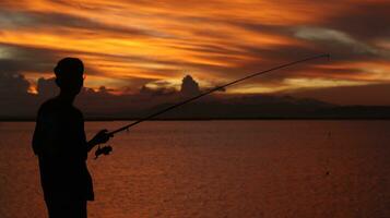 Silhouette von ein Fischer mit ein Angeln Stange im das See beim Sonnenuntergang foto