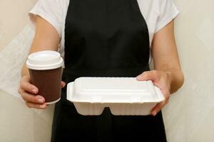 ein Frau Cafe Arbeiter dient ein abgeschlossen wegbringen bestellen ein Glas von Kaffee und ein Container mit Essen im Einweg Utensilien gemacht von recycelt roh Materialien foto