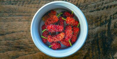 oben Aussicht von hell rot Maulbeere Obst im ein Schüssel auf hölzern Hintergrund, gesund frisch Maulbeere Obst foto