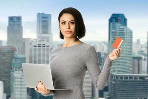Porträt von zuversichtlich Geschäftsfrau halt Laptop und Bank Karte im ihr Hände Über Stadt Hintergrund foto