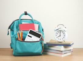 Vorderseite Aussicht von Grün Rucksack mit Schule liefern, Stapel von Bücher und Weiß Jahrgang Alarm Uhr 8 Uhr auf hölzern Tabelle und Weiß Hintergrund mit Kopieren Raum. foto