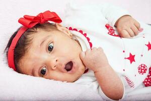 Nahansicht von ein Süss einer Monat Baby Mädchen mit ein Überraschung Ausdruck tragen ein rot und Weiß t Shirt. foto