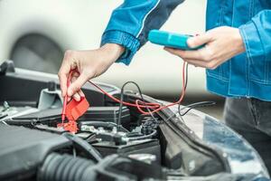 Auto Elektriker oder Mechaniker prüft Stromspannung im Auto Batterie Innerhalb das Auto Motor mit öffnen Kapuze foto
