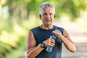 älter Mann tragen Kopfhörer öffnet oben ein Flasche von Wasser draußen nach das Cardio trainieren foto