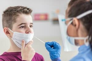 jung Junge tragen Gesicht Maske Über Mund hält immer noch wie Krankenschwester nimmt ein Stichprobe von seine Nase mit Tupfer foto