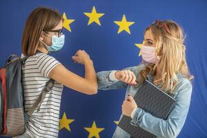 Ellbogen stoßen Gruß durch zwei weiblich Studenten tragen schützend Masken mit EU Flagge im das Hintergrund foto