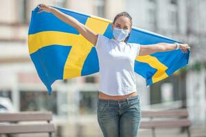 Schwedisch Mädchen auf ein Straße hält Flagge im ihr Hände Feiern, tragen ein Gesicht Maske foto