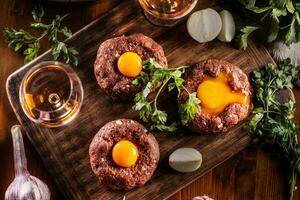 oben Aussicht von appetitlich Rindfleisch Steak Tartar mit köstlich Ei Eigelb und Alkohol. serviert auf ein hölzern Schreibtisch mit Zwiebel und Kräuter foto